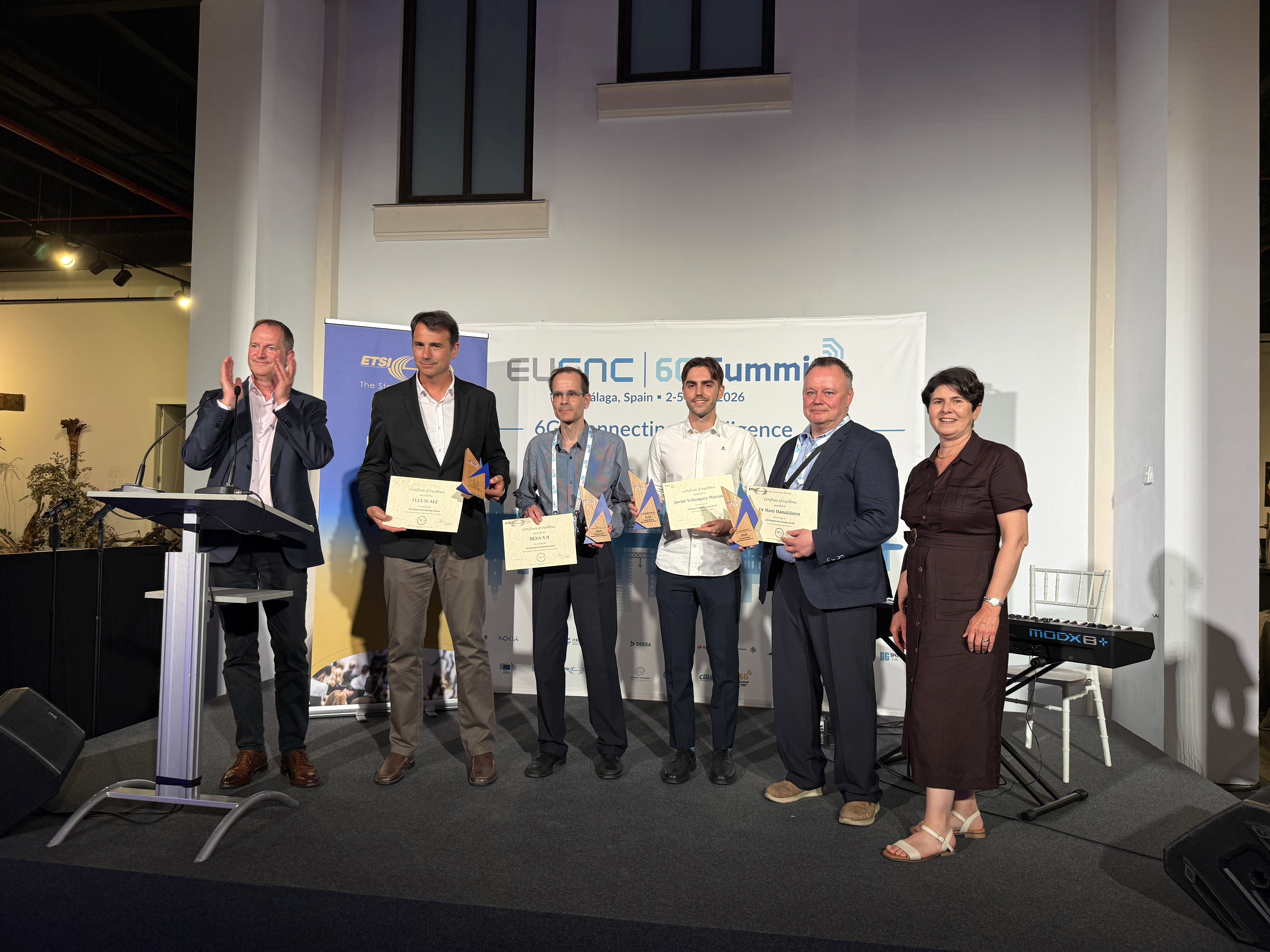 A group of six adults stand on a small indoor stage during an awards presentation. Five individuals in the center hold certificates and triangular trophies, while a person at a podium on the far left appears to be applauding. Behind the group is a large banner that reads “EuCNC & 6G Summit, Málaga, Spain – 2–5 June 2026,” along with additional text including “6G connecting intelligence.” An ETSI-branded banner stands to the left side of the stage.