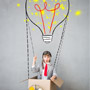 A child in an aviator hat sits in a cardboard box with a light bulb glowing above his head