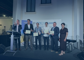 A group of six adults stand on a small indoor stage during an awards presentation. Five individuals in the center hold certificates and triangular trophies, while a person at a podium on the far left appears to be applauding. Behind the group is a large banner that reads “EuCNC & 6G Summit, Málaga, Spain – 2–5 June 2026,” along with additional text including “6G connecting intelligence.” An ETSI-branded banner stands to the left side of the stage.