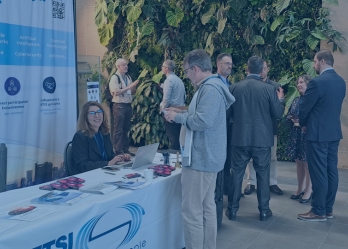 A networking scene at an indoor event space featuring an ETSI information stand on the left, where a seated staff member works on a laptop behind a table covered with brochures, flyers, and small promotional items. A vertical ETSI banner beside the table displays topics such as mobile networks, artificial intelligence, data, cybersecurity, and quantum technologies, along with key principles like global reach, direct participation, and independence. In the center and right of the image, a small group of attendees stand in conversation, wearing business or smart-casual attire. The background features a large green living plant wall, adding a natural element to the space, while the right side includes a tall abstract wooden sculpture mounted in a rectangular planter. The setting appears to be a modern conference or networking venue with tiled flooring and soft indoor lighting.