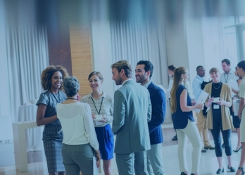 A group of event participants wearing conference badges stand in small groups, talking and networking in a bright indoor venue during a professional industry event.